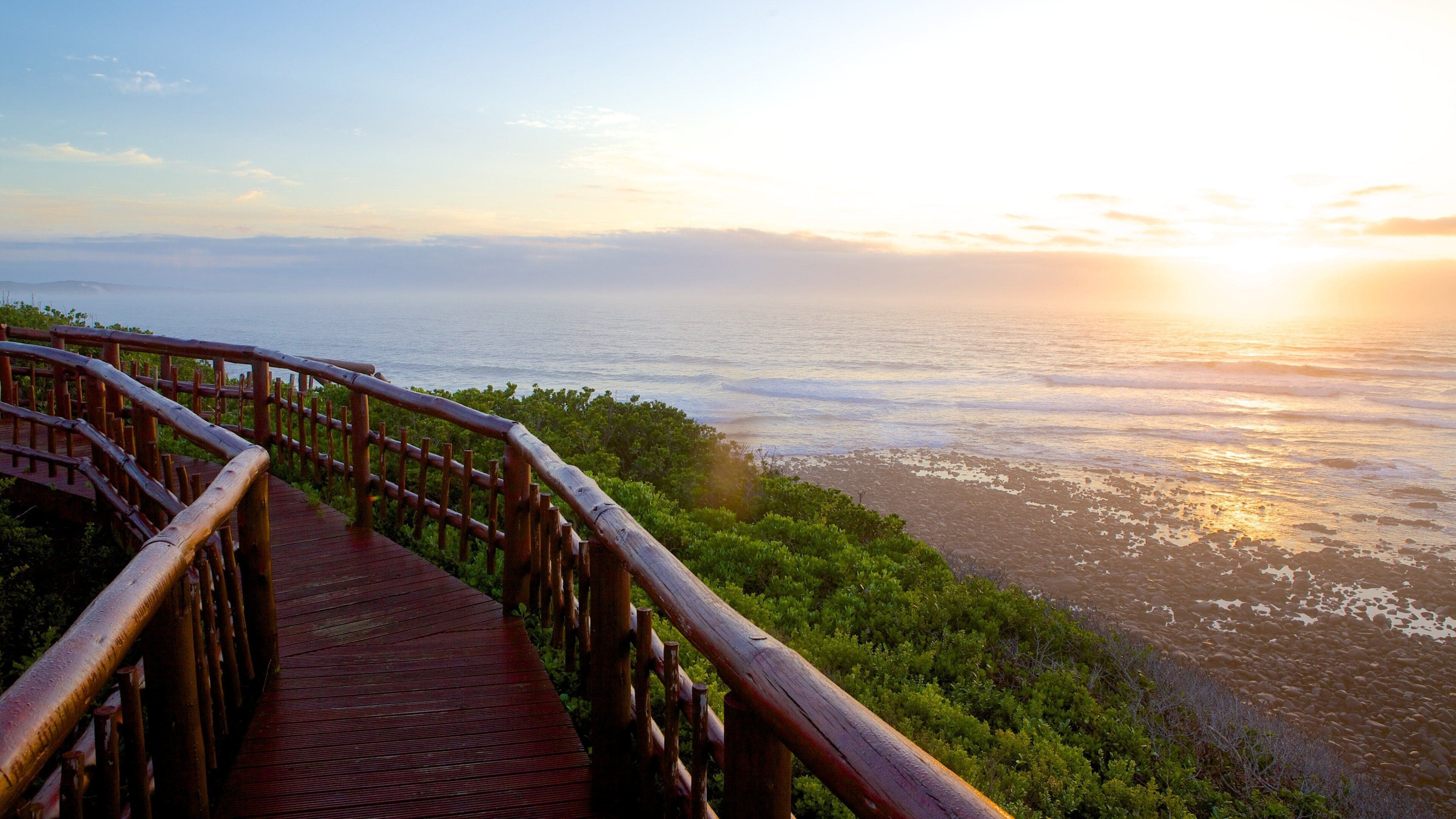 A wooden boardwalk leading to a coastal view with the ocean and a sunset in the background.