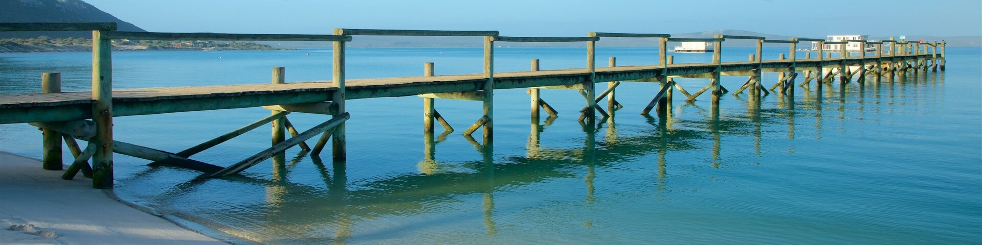 Eine Holzsteg, der sich in ruhiges Wasser erstreckt, wo sich die Umgebung deutlich spiegelt, ein Sandstrand und ein ferner Berg.