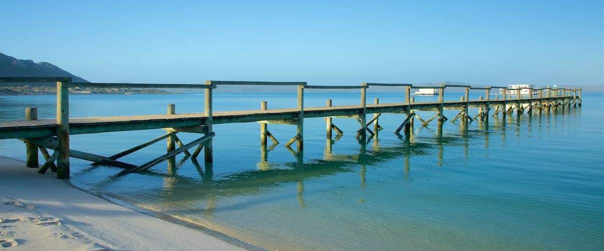 A wooden pier extending into calm waters with clear reflections, a sandy beach, and a distant mountain.