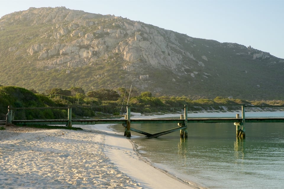 A beach with a wooden pier leading to a calm body of water, surrounded by a rocky mountain and greenery.