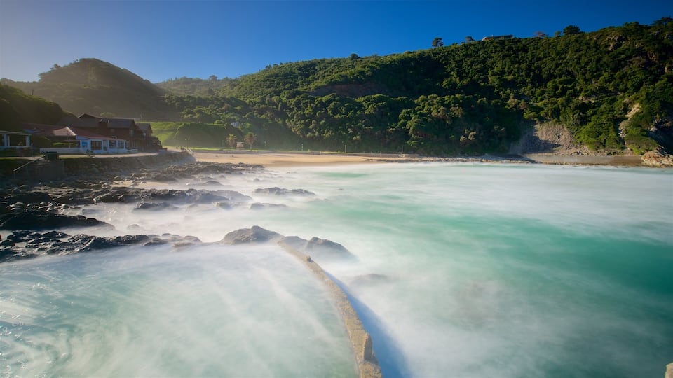 A coastal landscape with a rocky shoreline, clear turquoise waters, and a forested hill in the background.