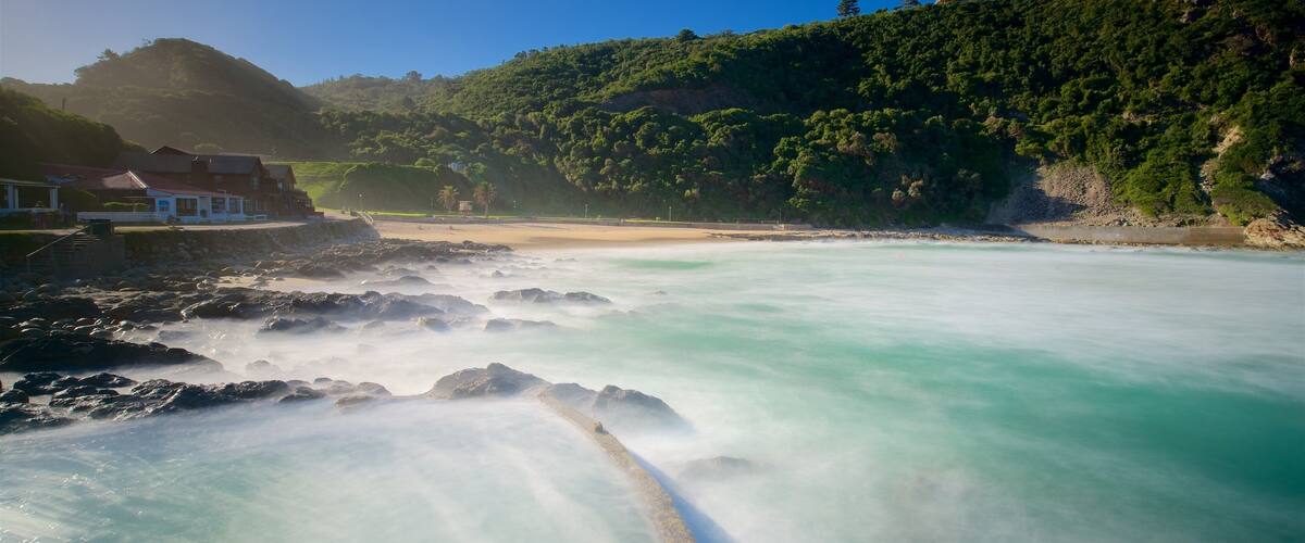 A coastal landscape with a rocky shoreline, clear turquoise waters, and a forested hill in the background.