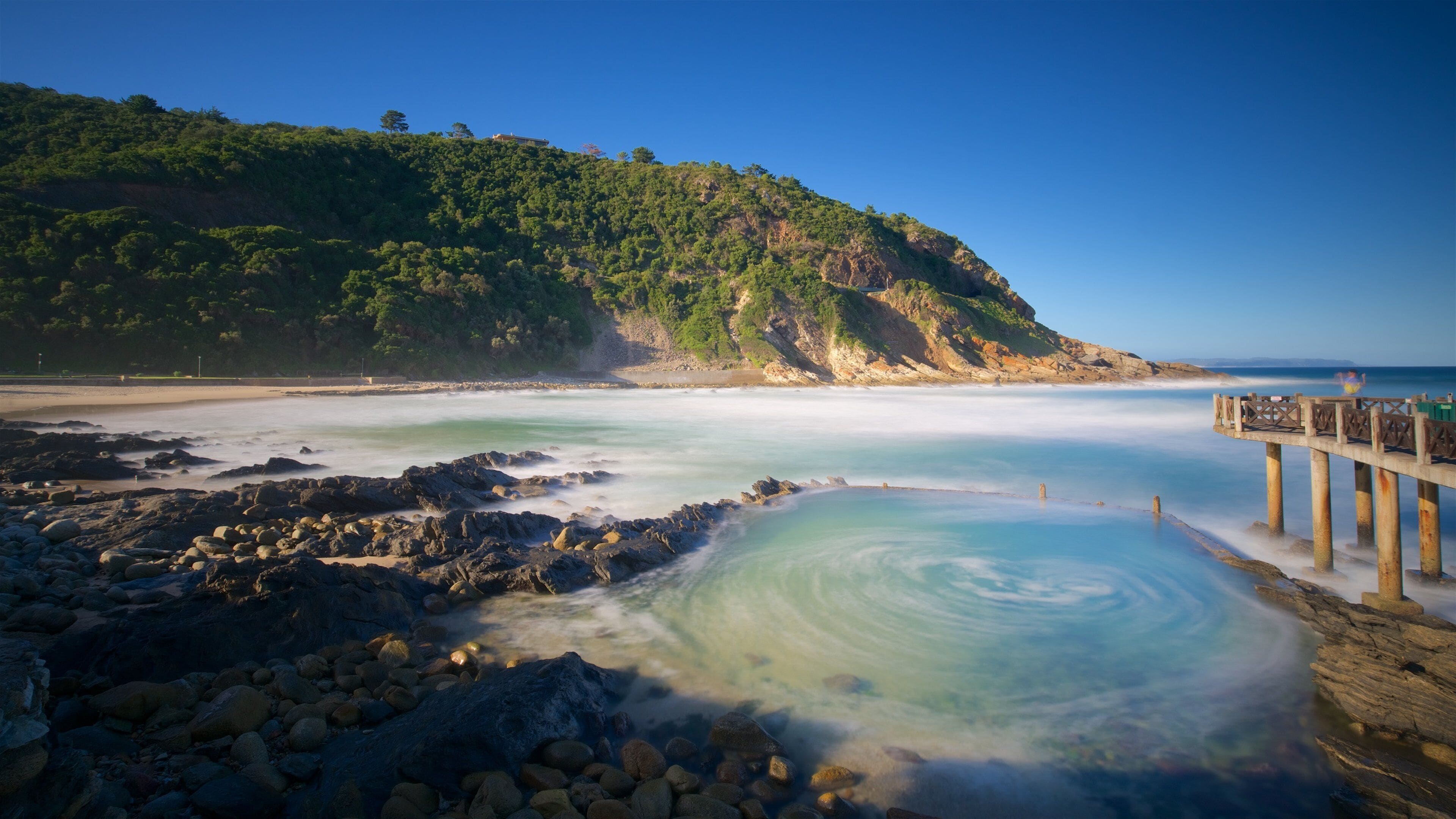 A coastal landscape with a rocky shoreline, a wooden pier, and a forested hill in the background.