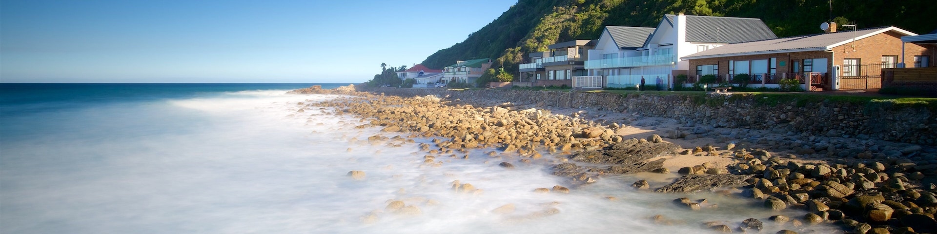 Una playa con costa rocosa, casas y montañas al fondo.