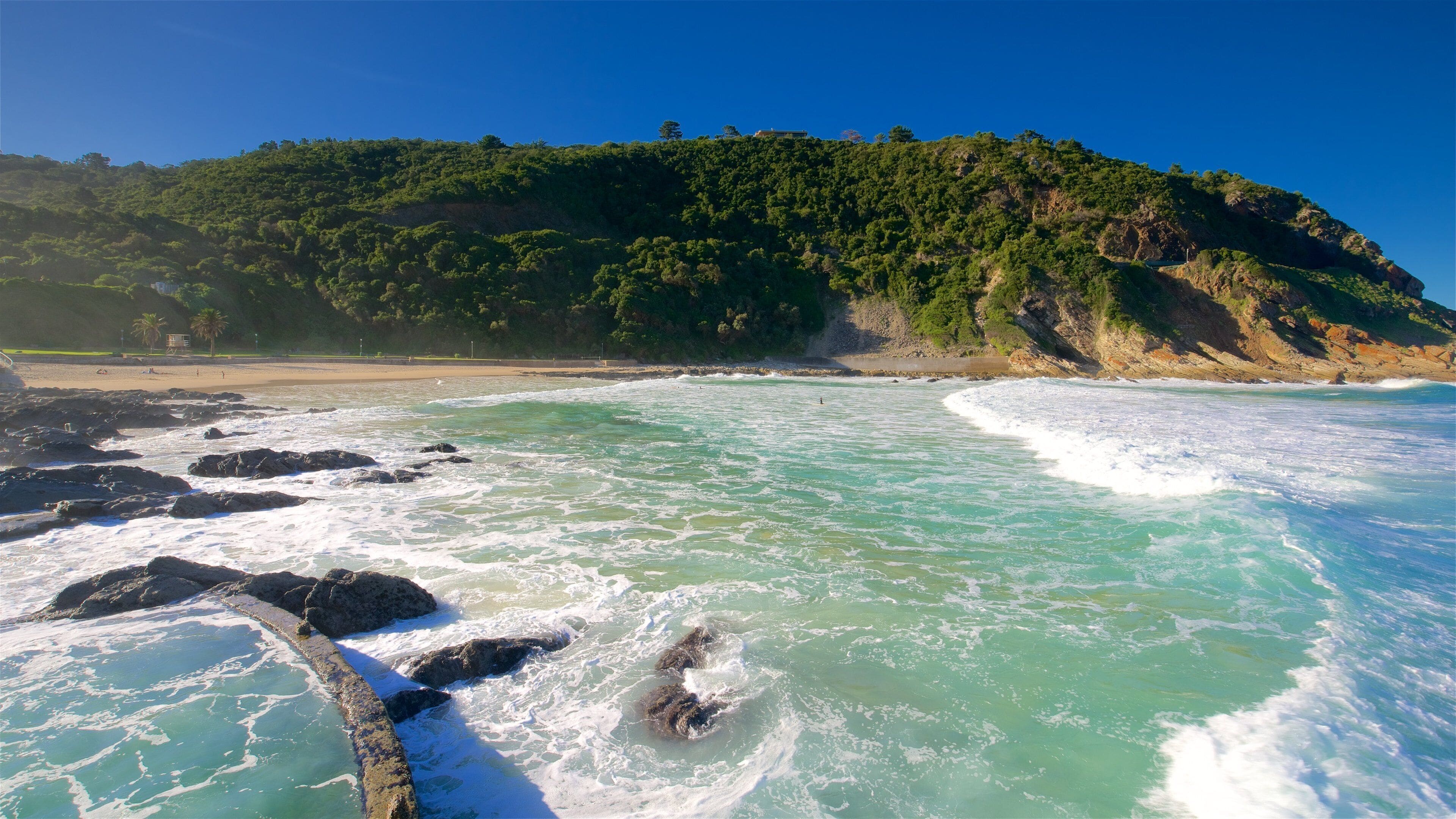 Ein Strand mit türkisblauem Wasser, felsigem Ufer und einem bewaldeten Hügel im Hintergrund.