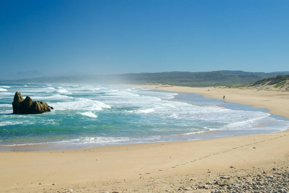 Platja sorrenca amb ones que trenquen a la riba, un promontori rocos on brollen les onades i una persona fent de pesca.