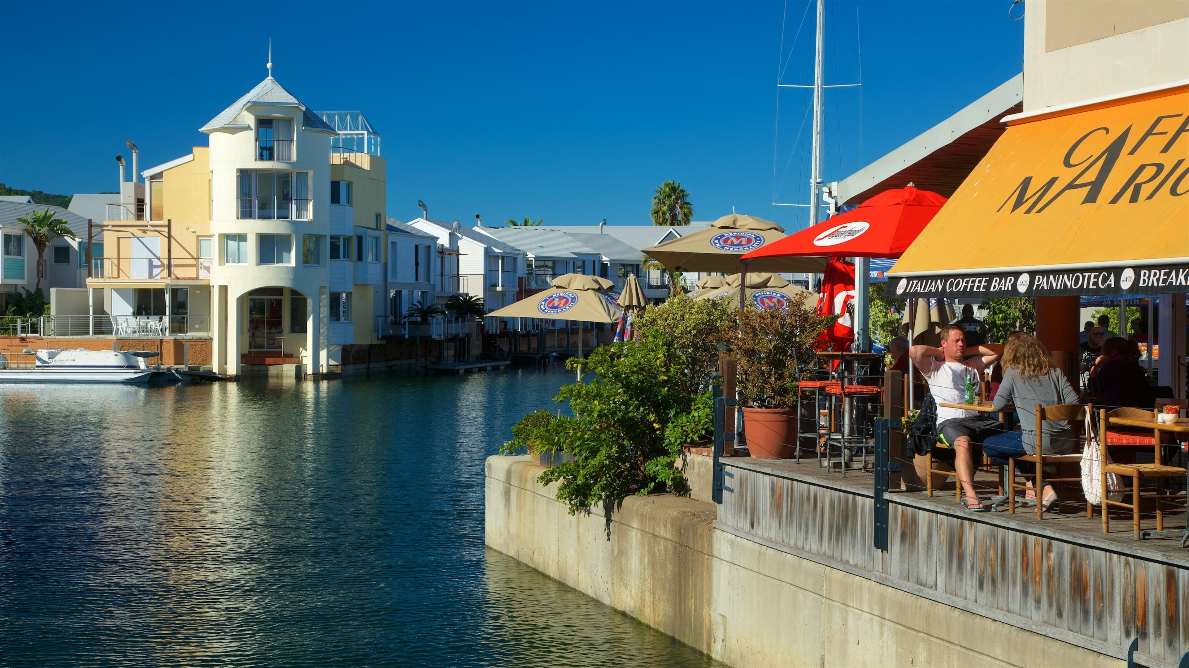 A waterfront café with outdoor seating, a canal, and a building with a tower.