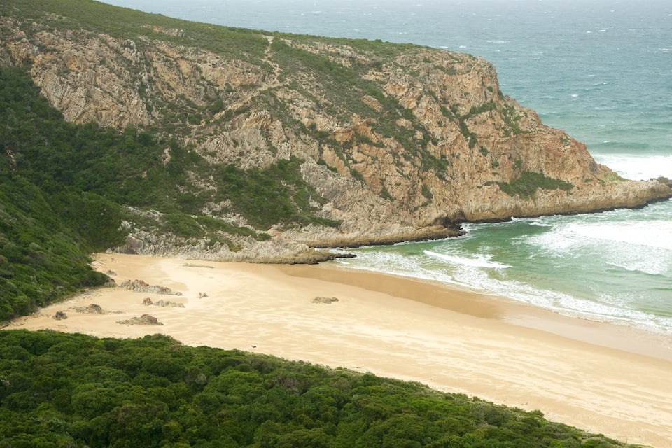 A coastal landscape with a sandy beach, rocky cliffs, and dense greenery.