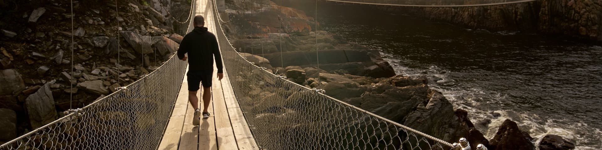 A person walking on a suspension bridge over a river with rocky cliffs on either side.