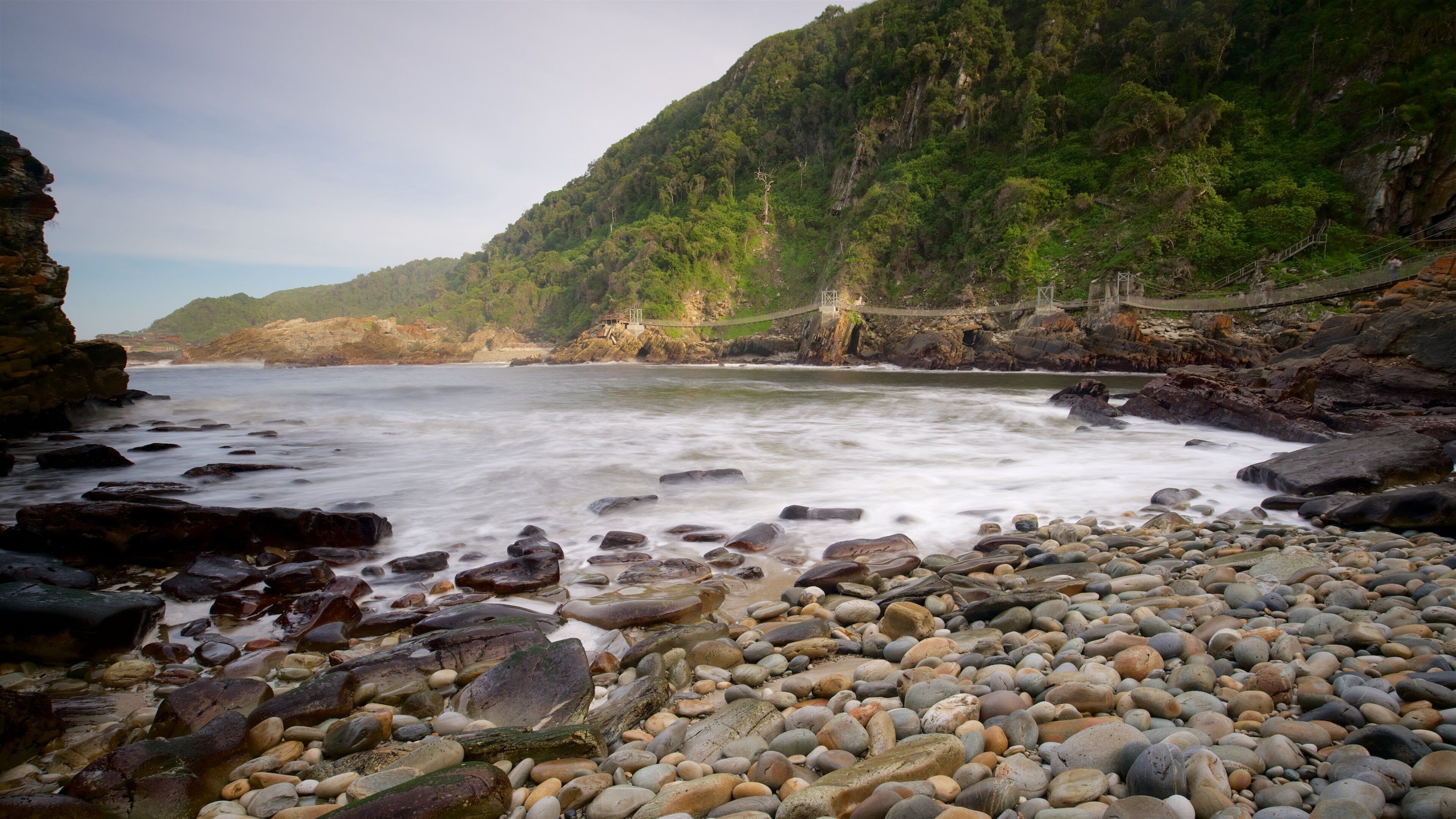 A rocky beach with a suspension bridge and lush green hills in the background.