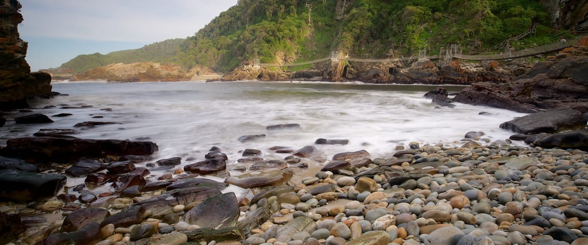 A rocky beach with a suspension bridge and lush green hills in the background.