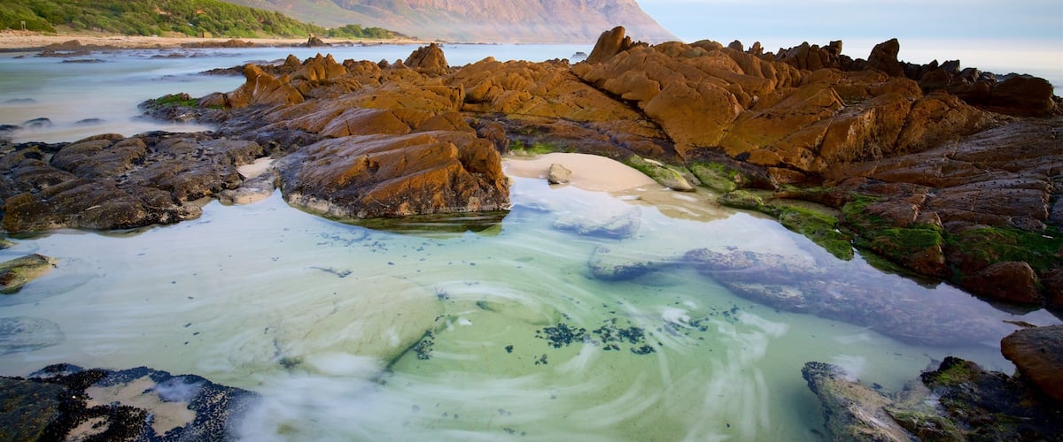 Eine Küstenlandschaft mit Felsformationen, klarem, flachem Wasser und entfernten Bergen.