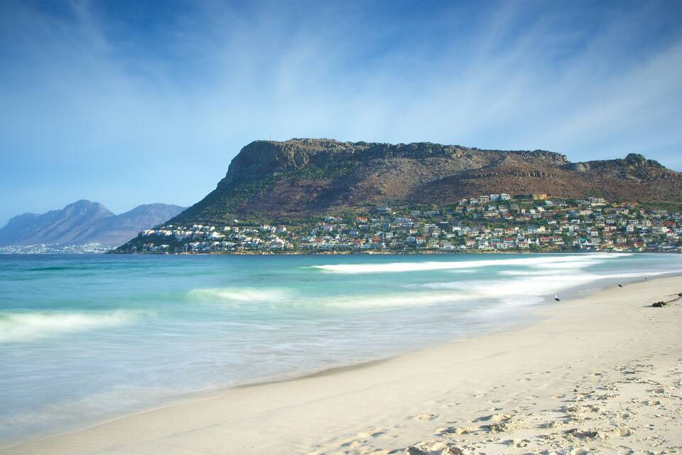 A beach with clear turquoise waters, a sandy shore, and a mountain with a town on its slopes.