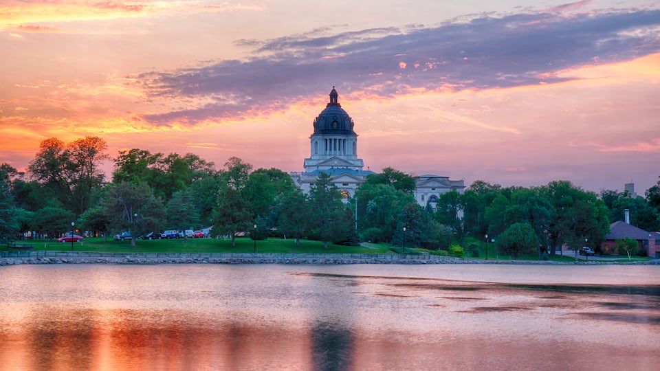 A large domed building with a reflection in a calm body of water, surrounded by trees and a sunset sky.