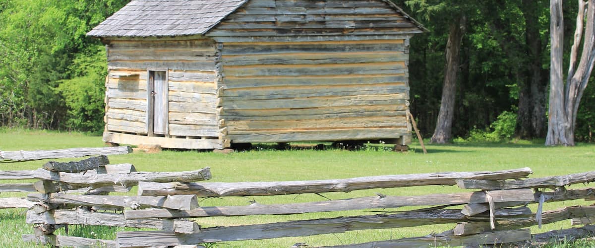 A small wooden cabin with a sloped roof, surrounded by a grassy field and trees.