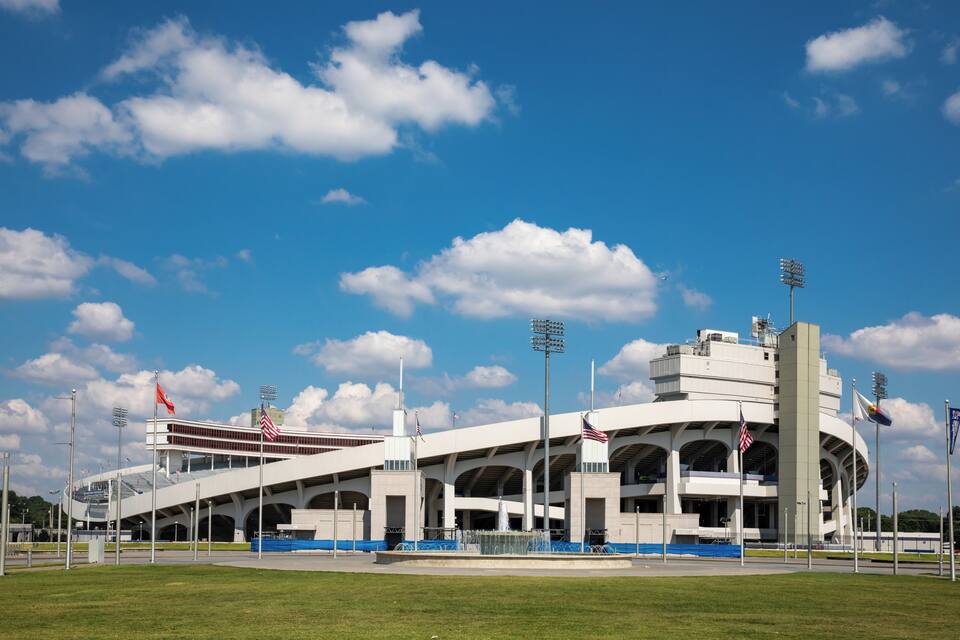 A stadium with a white exterior, multiple flagpoles, and a clear blue sky with clouds.
