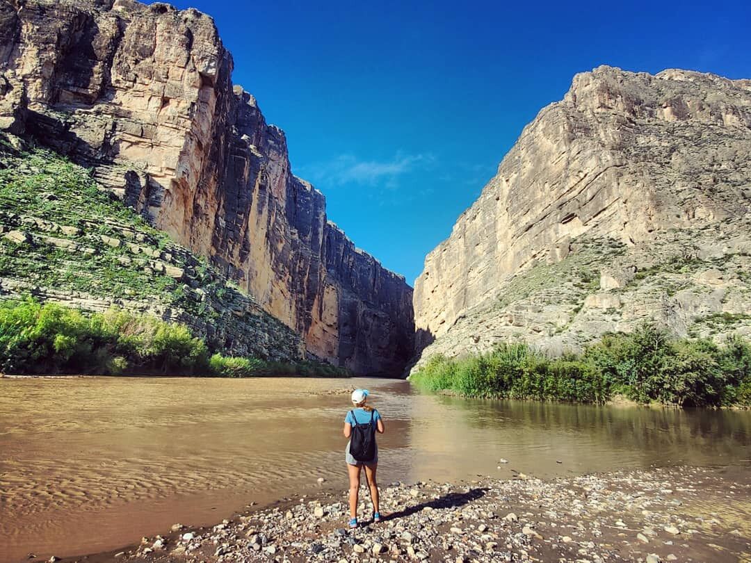 A person standing by a river with rocky banks and steep canyon walls.