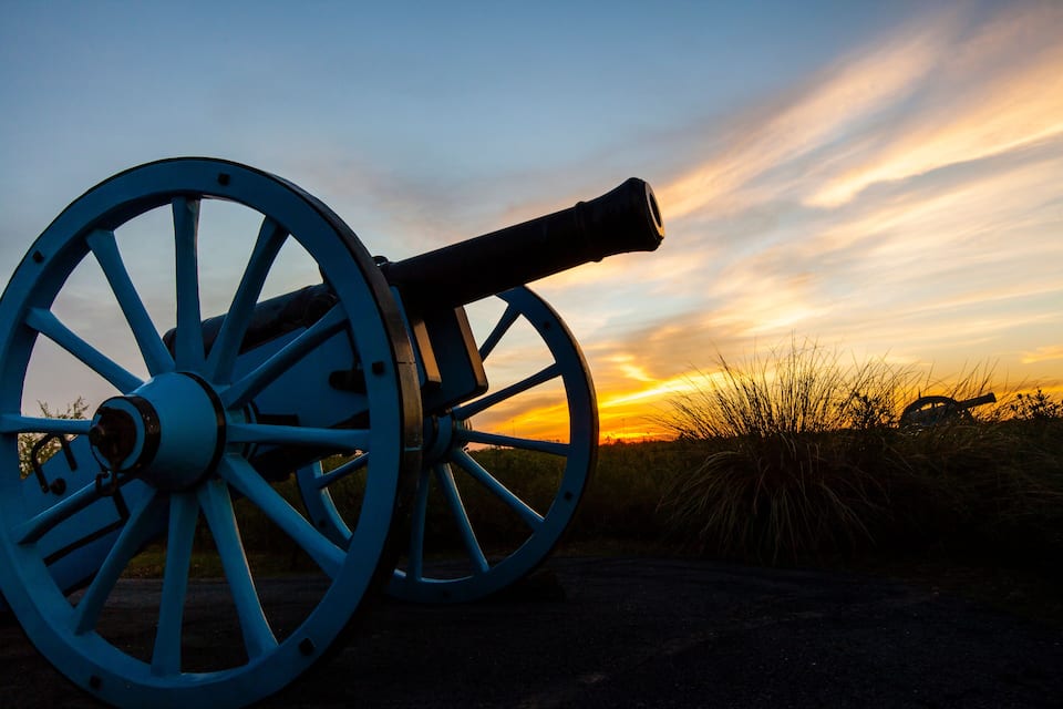 A cannon with large blue wheels against a sunset sky.