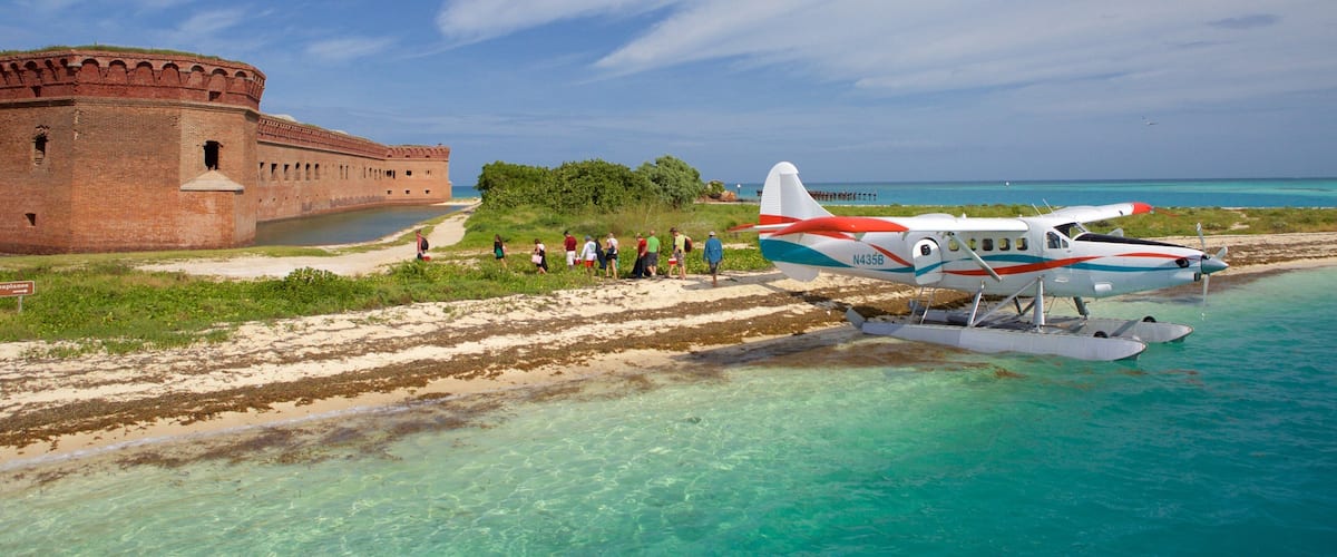 A beach with a historic brick fort, a small seaplane, and a group of people walking along the shore.