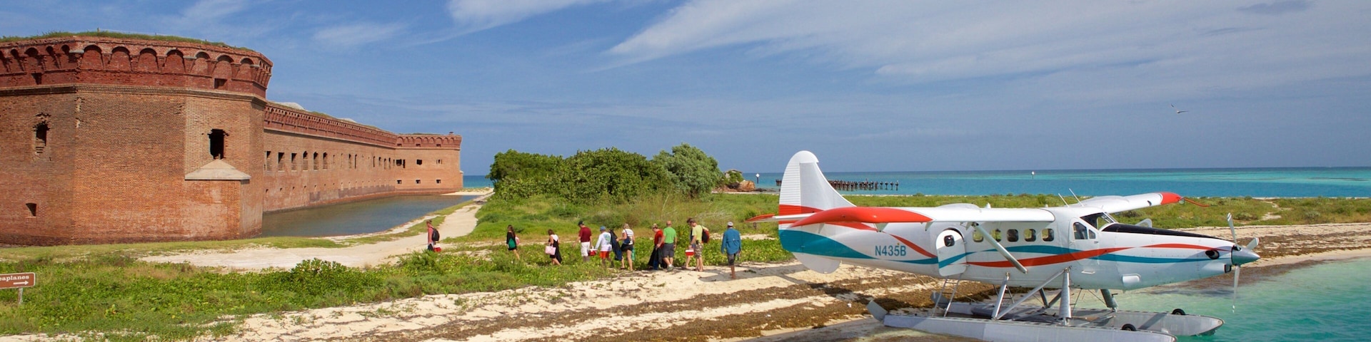 Una playa con una antigua fortaleza de ladrillo, un pequeño hidroavión y un grupo de personas caminando por la orilla.