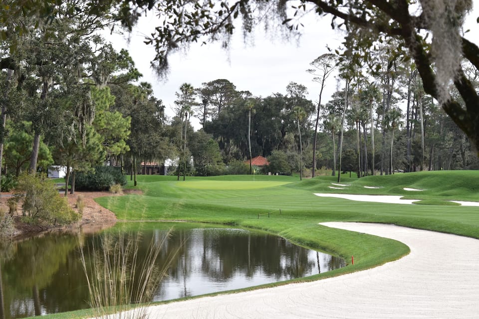 Een golfbaan met een vijver, zandbunkers en een clubhuis op de achtergrond.