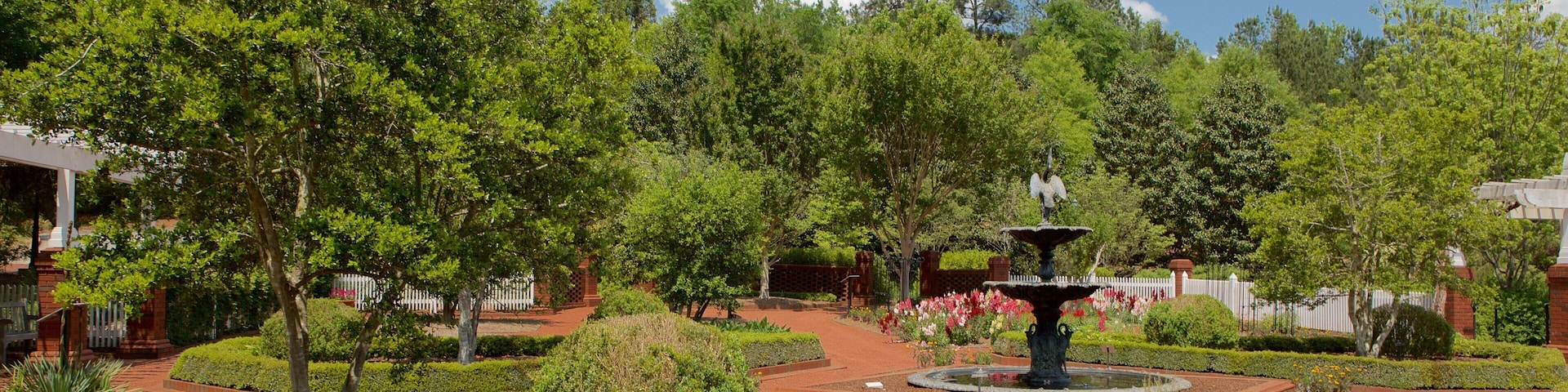 Um jardim paisagístico com uma fonte central, rodeado por sebes e árvores bem cuidadas.