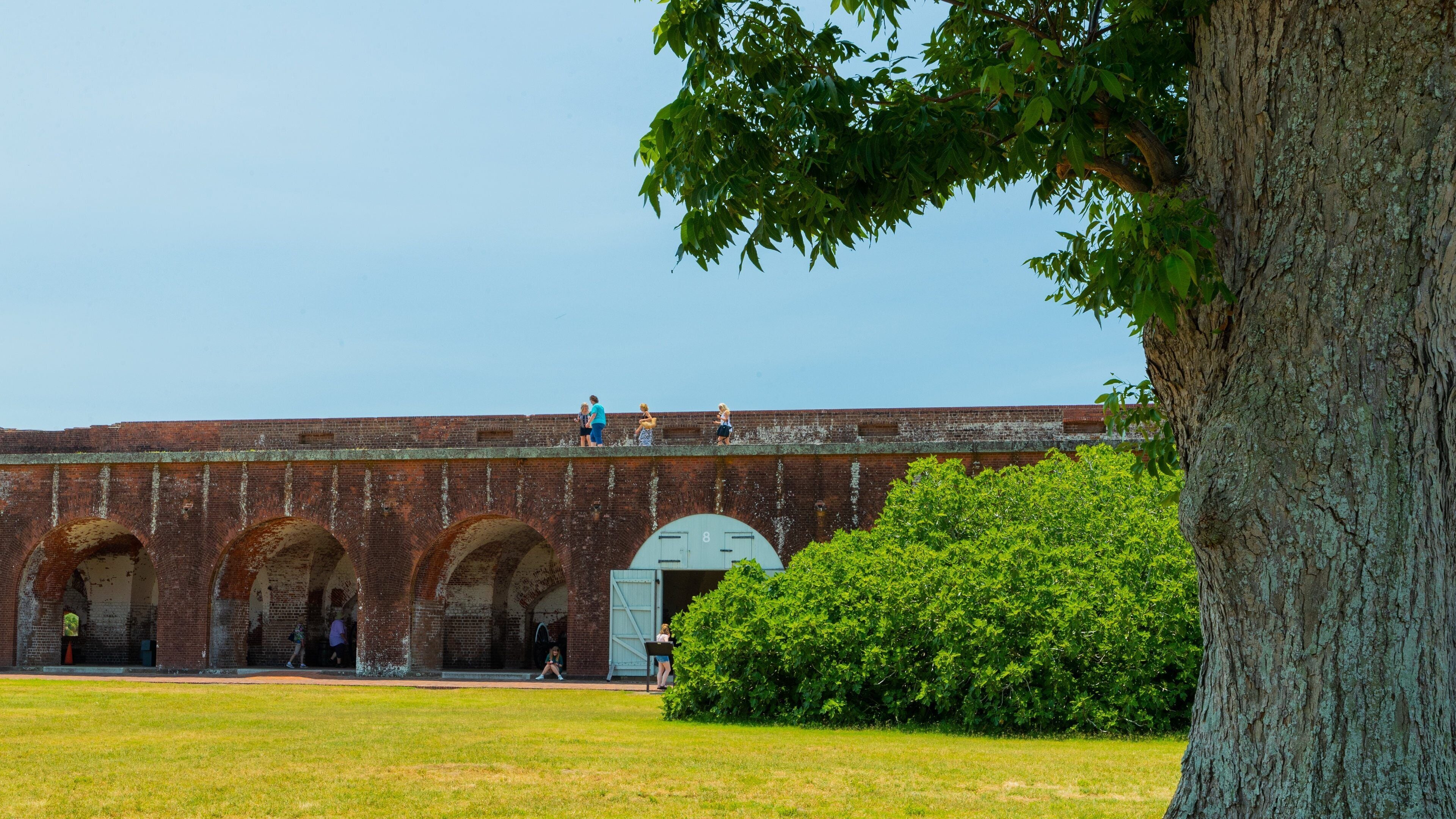 A brick building with arched doorways and a large tree in the foreground.