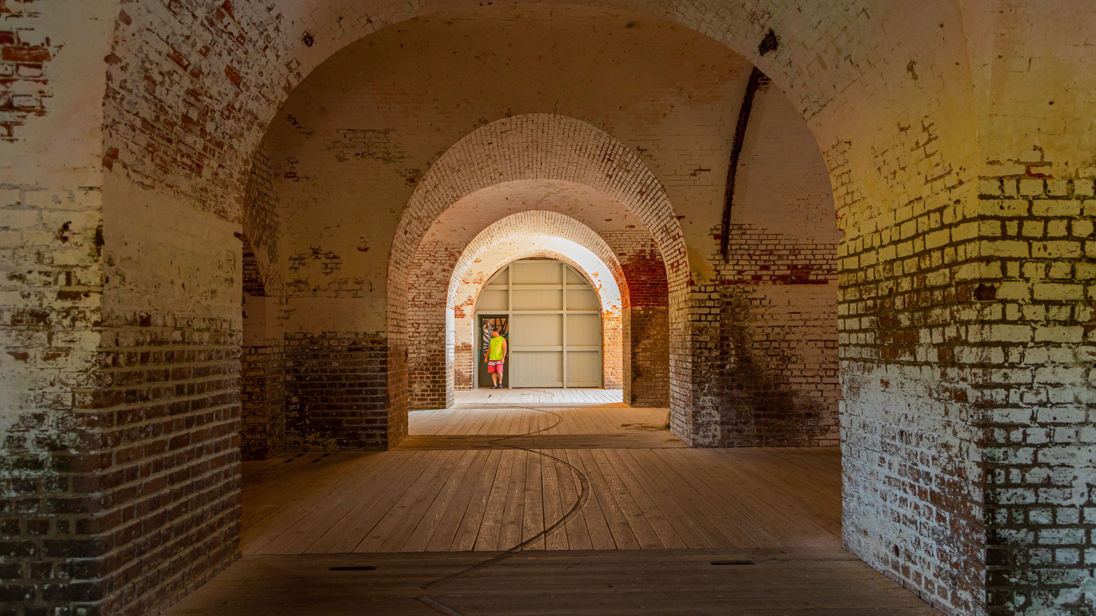 An arched brick corridor with a wooden floor and a person in a yellow vest standing near a door.