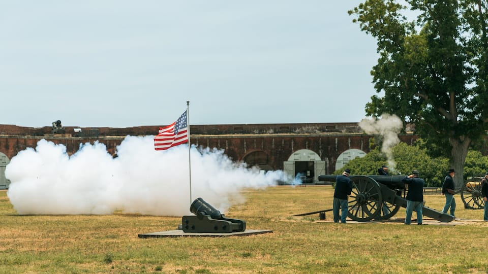 A cannon firing with smoke, an American flag, and a historic fort in the background.