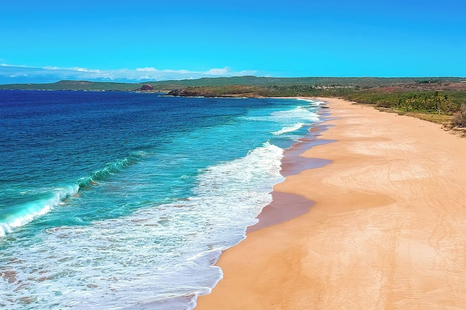 A beach with clear turquoise water, sandy shore, and distant green hills.