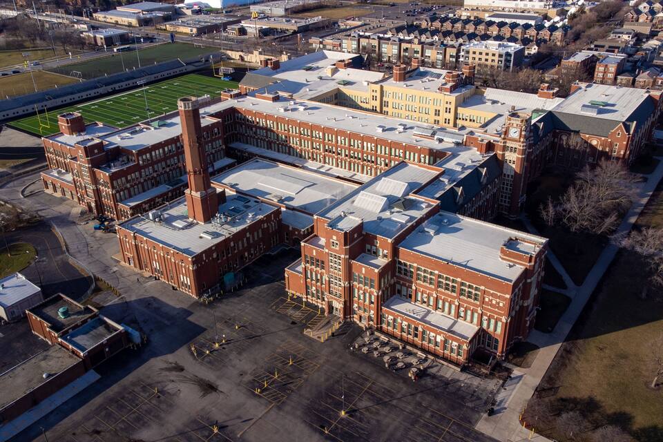 An aerial view of a large industrial complex with multiple buildings, a football field, and a parking lot.