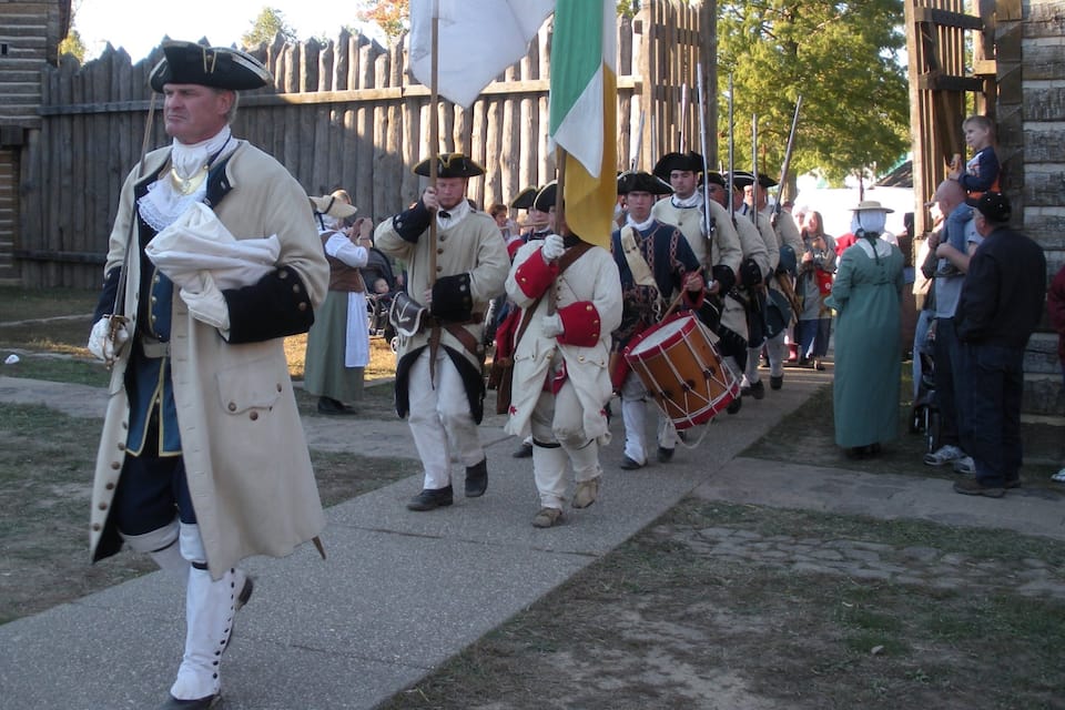 A historical reenactment with participants in period costumes and uniforms marching in formation.