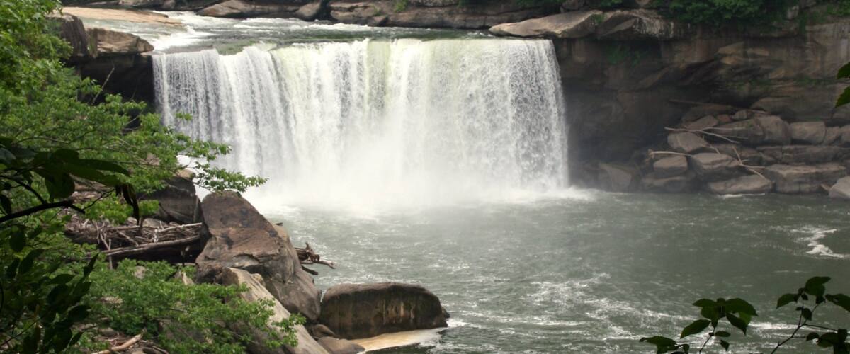 A waterfall cascading over rocks into a river, surrounded by dense forest.