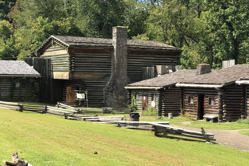 A rustic log cabin with a stone chimney, surrounded by a wooden fence and a grassy field.