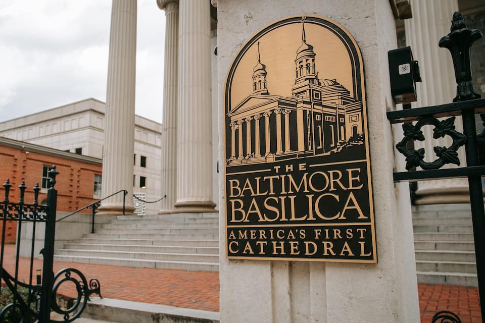 Un letrero de la Basílica de Baltimore, la primera catedral de Estados Unidos, con columnas de estilo clásico y una ilustración detallada del edificio.