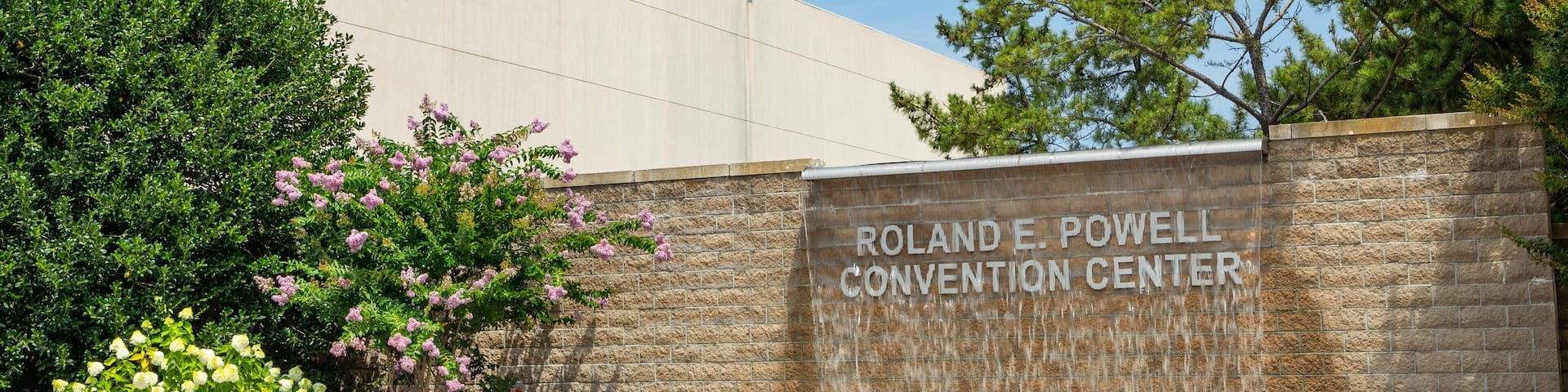 A stone wall with the text "Roland E. Powell Convention Center" and a landscaped area with trees and shrubs.