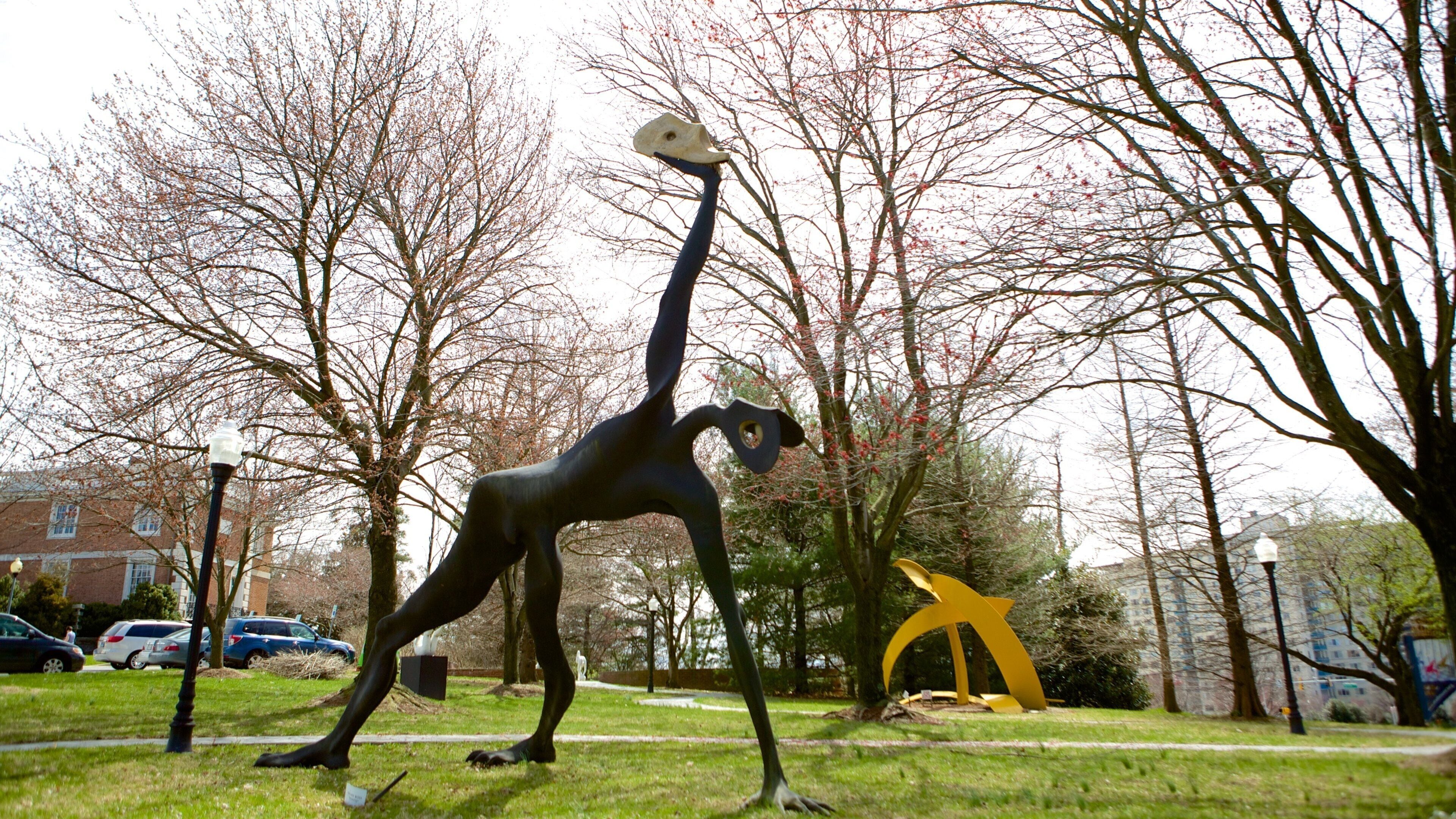 A sculpture of a human figure in a park with trees and a yellow abstract sculpture in the background.