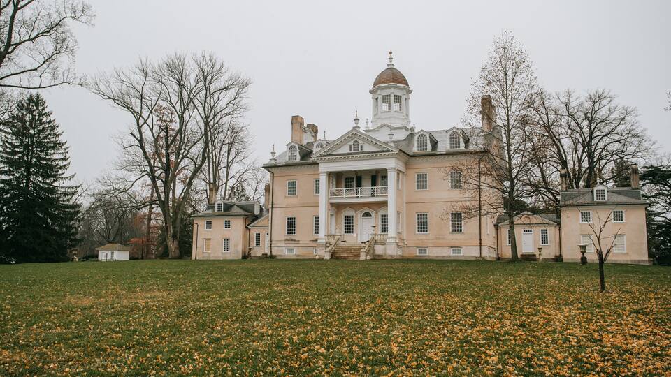 A large, historic mansion with a prominent tower, surrounded by a grassy field and leaf-covered ground.