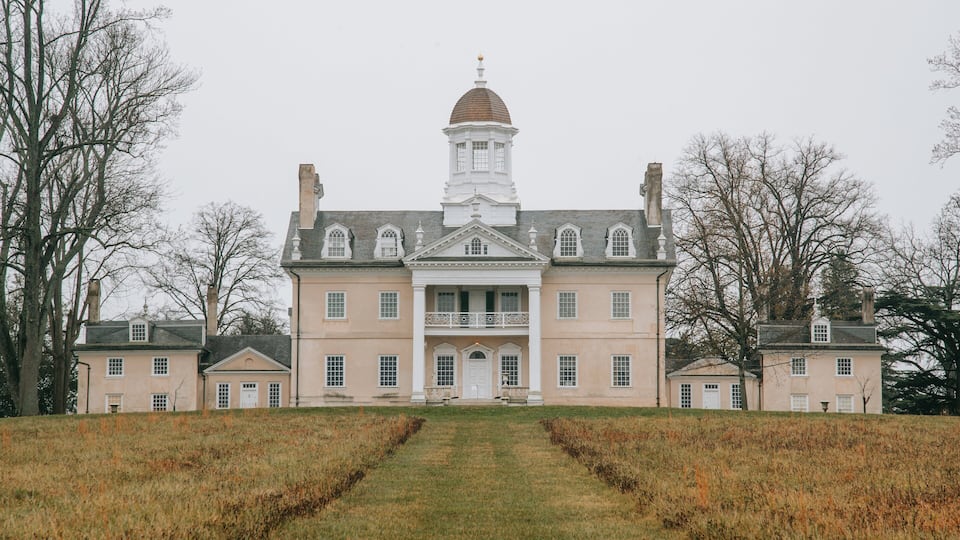 A large stately building with a central dome and symmetrical wings, surrounded by a grassy field and trees.