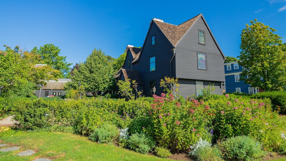 A large, dark-colored house with a steeply pitched roof, surrounded by lush greenery and flowering plants.