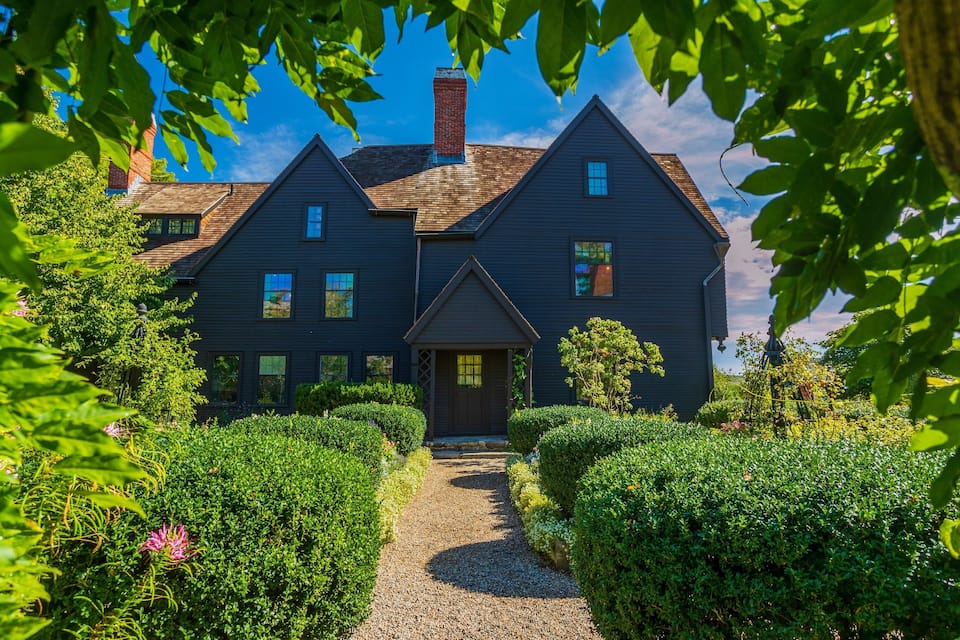 A dark wooden house with a chimney, surrounded by lush greenery and a gravel path.