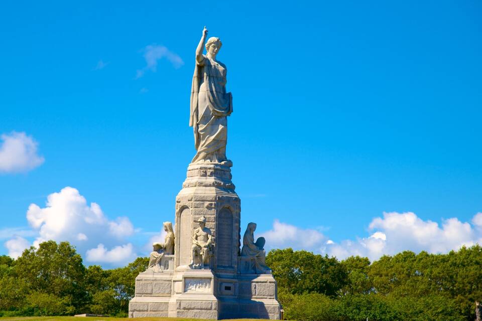 A large stone statue of a woman holding a torch, surrounded by smaller statues and set against a clear blue sky.