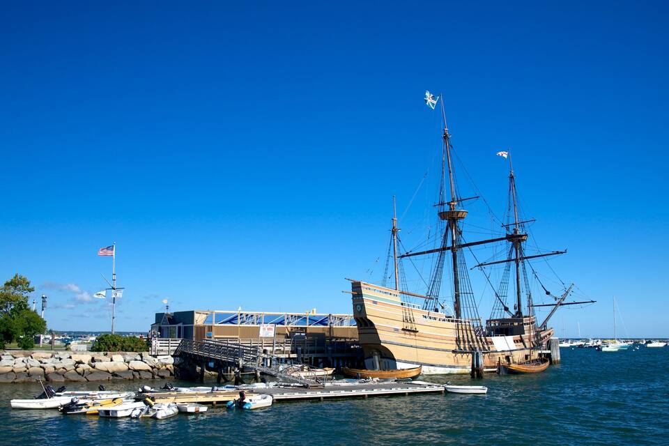Een historisch schip afgemeerd aan een steiger, met verschillende kleinere boten in de buurt.