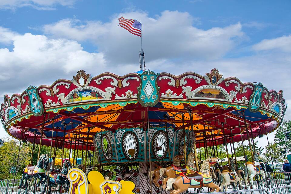 Un carrusel colorido con una bandera estadounidense en la parte superior, rodeado de decoraciones elaboradas y un cielo azul despejado.
