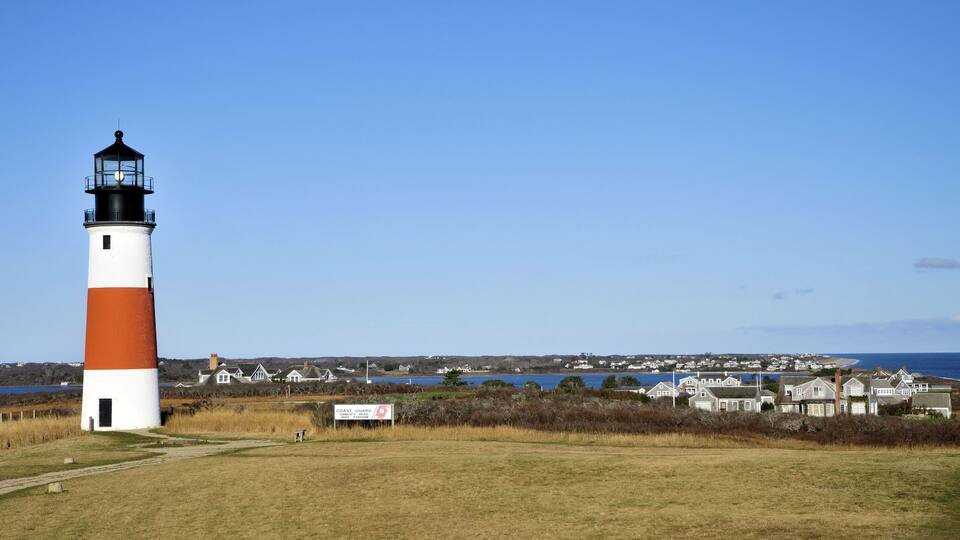 A lighthouse with a white and orange color scheme, a sign indicating it is a Coast Guard station, and a coastal town in the background.