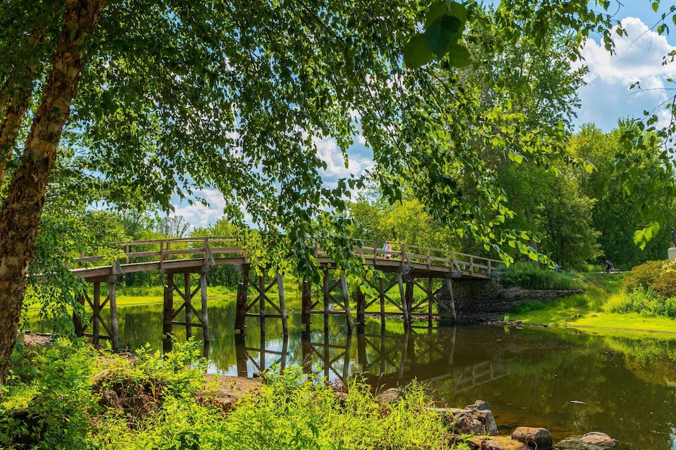 Eine Holzbrücke über einem ruhigen Fluss, umgeben von üppiger Vegetation und einer Statue auf einem Hügel.