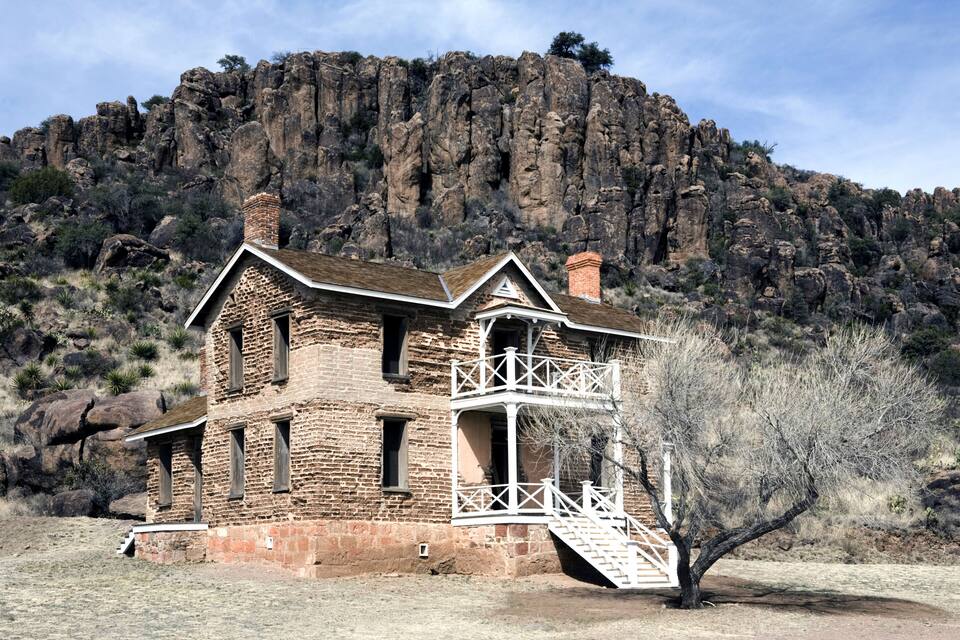 Una casa de piedra rústica con un balcón, frente a imponentes riscos y vegetación escasa.