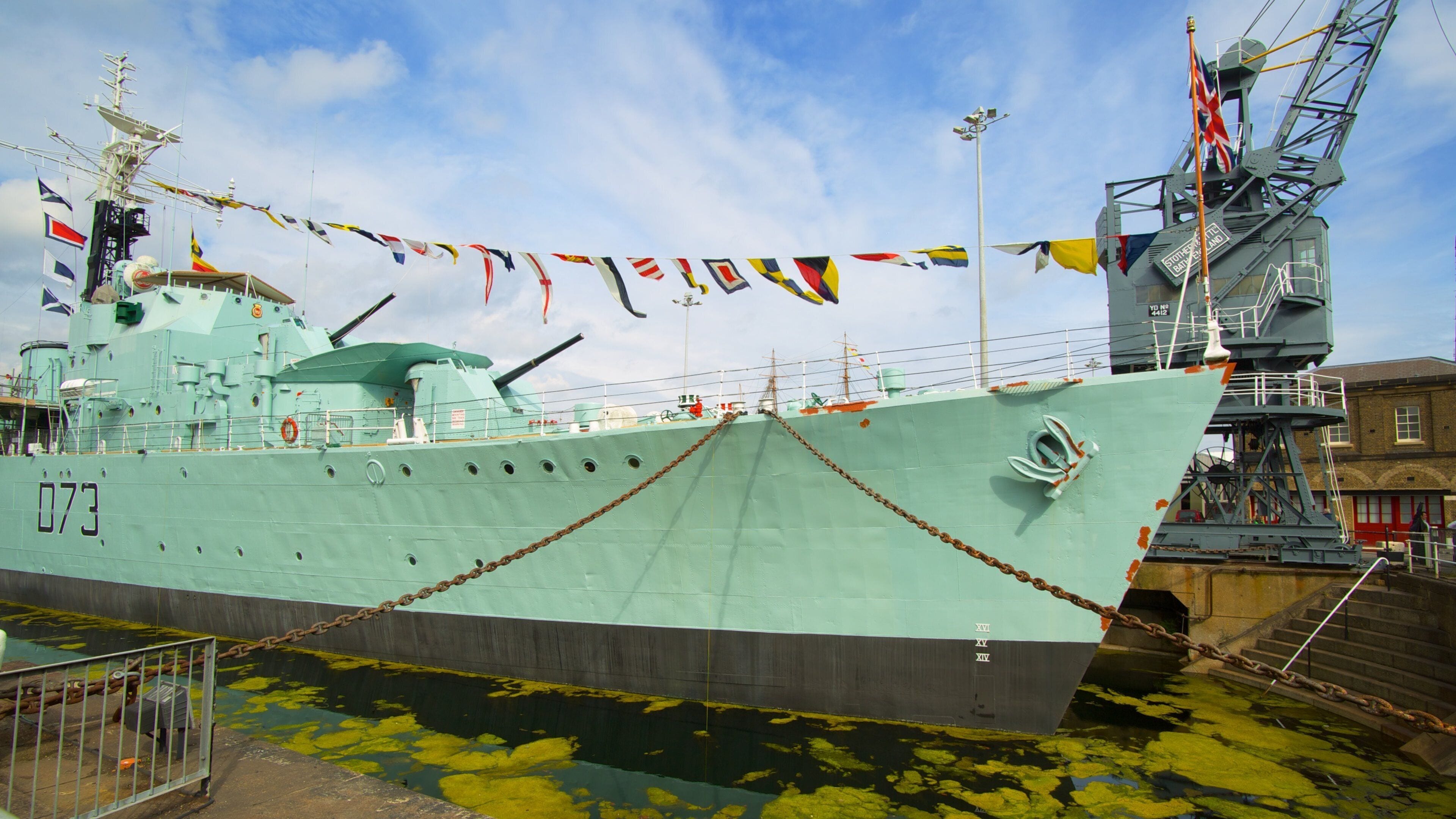 A large naval ship docked at a port with multiple flags and a crane structure nearby.