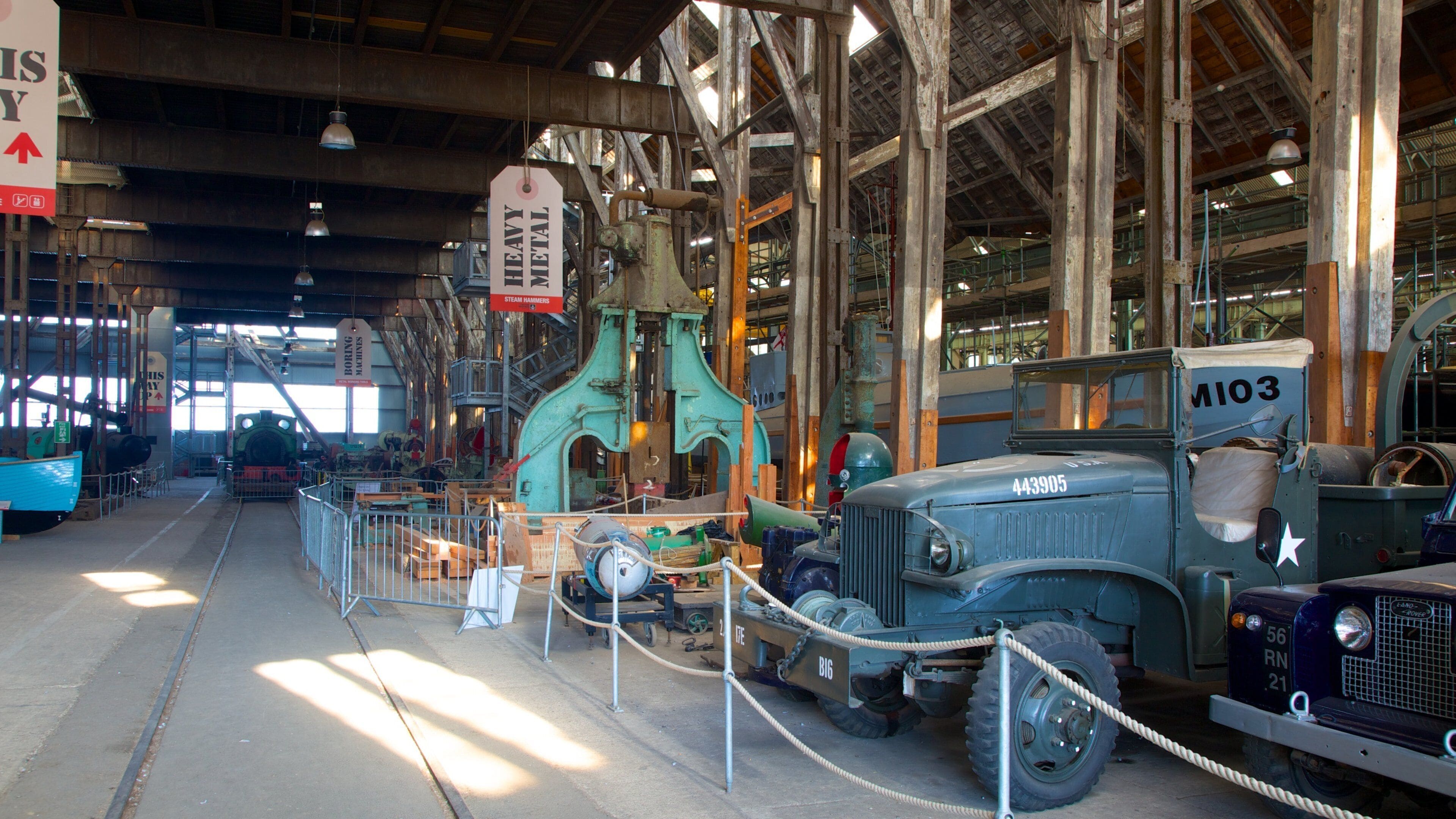 An industrial setting with vintage machinery, including a steam hammer and a large green machine, surrounded by metal railings and ropes.