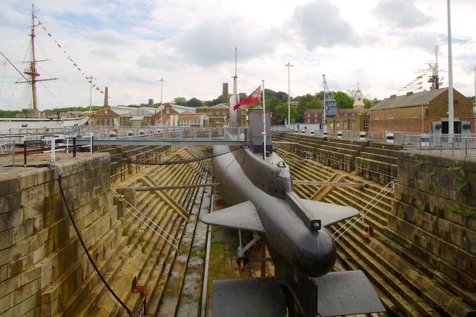 A submarine on display in a dock with a red flag, surrounded by buildings and a cloudy sky.